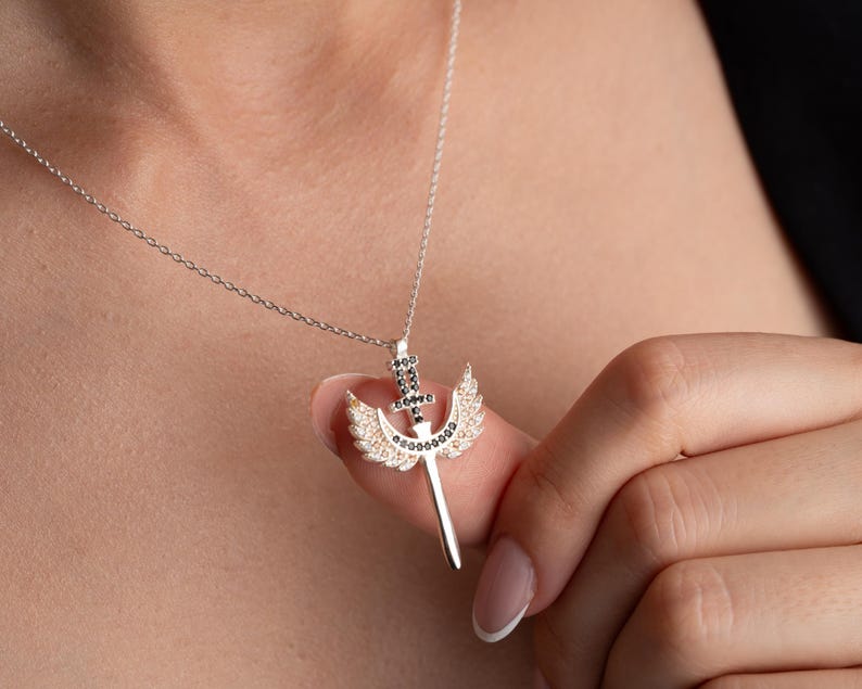 Puede incluir: Un collar de plata con un colgante que representa una espada con alas. La espada y las alas est&aacute;n adornadas con peque&ntilde;as piedras negras y blancas.