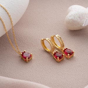 Könnte beinhalten: Ein goldener Halsketten- und Ohrring-Set mit roten tropfenförmigen Steinen und kleinen klaren Steinen. Der Schmuck befindet sich auf einer hellrosa Oberfläche.