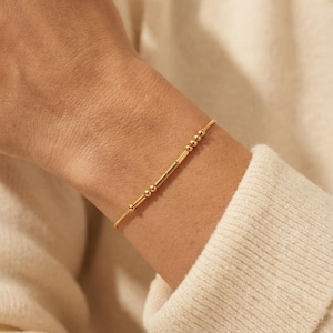 Könnte beinhalten: Ein zartes goldenes Armband mit einer Reihe kleiner, runder Perlen und rechteckiger Stäbe. Das Armband wird am Handgelenk getragen, wobei die Goldfarbe mit der Haut und einem cremefarbenen Pullover kontrastiert.