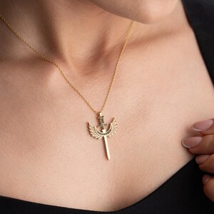 Puede incluir: Un collar de oro con un colgante que representa una espada con alas. El colgante est&aacute; adornado con peque&ntilde;as piedras negras.