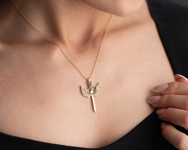 Puede incluir: Un collar de oro con un colgante que representa una espada con alas. El colgante est&aacute; adornado con peque&ntilde;as piedras negras.