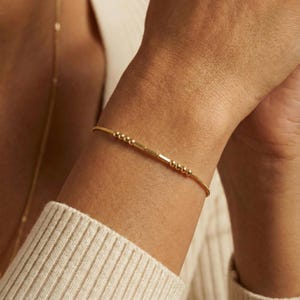 May include: A delicate gold bracelet featuring a series of small, round beads and rectangular bars. The bracelet is worn on a wrist, showcasing its minimalist design and subtle elegance. The bracelet's slender profile and warm gold tone suggest a refined aesthetic.