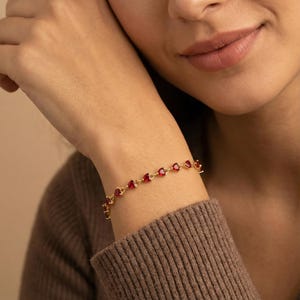 Può includere: Un braccialetto dorato con pietre preziose rosse a forma di cuore. Il braccialetto è indossato su un polso, con le pietre preziose uniformemente distanziate lungo la catena. Il gioiello è un accessorio delicato.