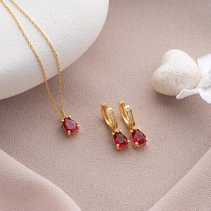 Może przedstawiać: Zestaw złotych kolczyków i naszyjnika z czerwonymi kamieniami w kształcie łzy. Kolczyki są w stylu koła, a naszyjnik ma delikatny łańcuszek.