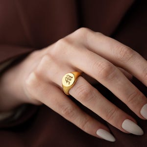 Puede incluir: Un anillo de sello de oro con un monograma personalizado. El anillo es de forma ovalada y presenta un diseño en relieve.