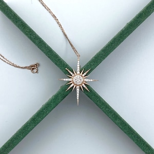 May include: A gold necklace with a starburst pendant set with clear cubic zirconia stones. The necklace is displayed on a green background.