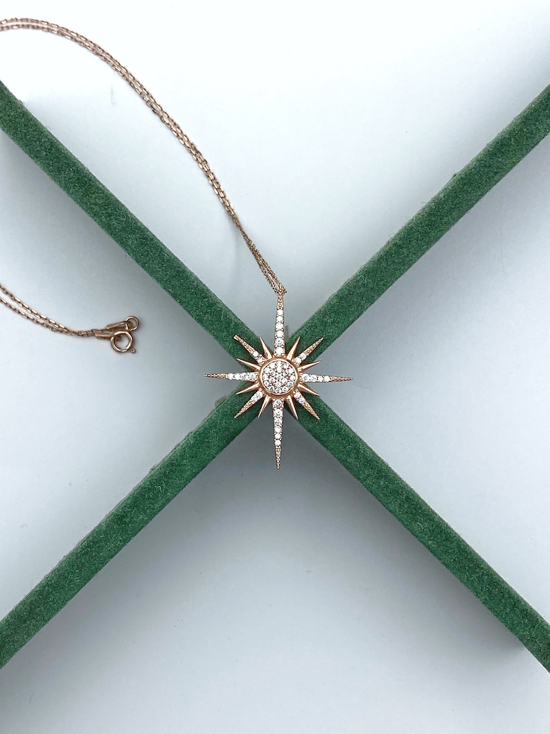 May include: A gold necklace with a starburst pendant set with clear cubic zirconia stones. The necklace is displayed on a green background.