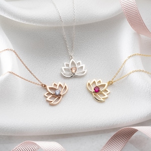 May include: Three necklaces with lotus flower pendants. One is rose gold with a light blue gem, one is silver with a peach gem, and the third is gold with a red gem. The necklaces are on a white, textured surface with a pink ribbon.