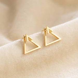 Puede incluir: Un par de pendientes de botón triangulares dorados. Cada pendiente presenta un diseño geométrico con un marco triangular y un pequeño detalle de flecha que apunta hacia arriba. Los pendientes se muestran sobre una superficie crema suave.
