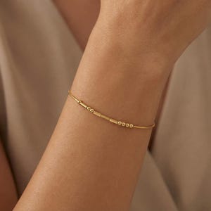 以下が含まれることがあります： 小さなビーズと長方形のバーが等間隔に並んだ繊細なゴールドブレスレット。細くてミニマリストなブレスレットはシンプルなデザインです。ブレスレットは人の手首に表示されています。