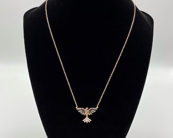 Pode incluir: Um colar de ouro rosa com uma corrente delicada e um pingente de pássaro. O pássaro é adornado com pedras pretas e brancas.