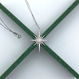 May include: A silver necklace with a starburst pendant set with clear cubic zirconia stones. The necklace is on a green velvet background.