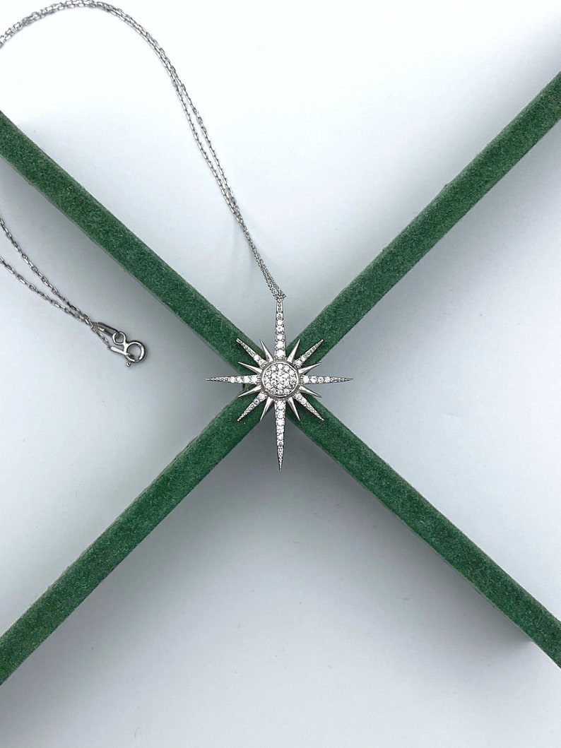May include: A silver necklace with a starburst pendant set with clear cubic zirconia stones. The necklace is on a green velvet background.