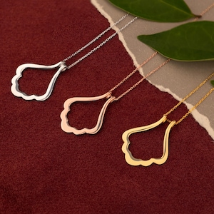 Puede incluir: Tres collares con colgantes en forma de lágrima con borde festoneado. Los colgantes son plateados, oro rosa y dorados, cada uno con una cadena delicada. El fondo es una superficie roja texturizada con papel rasgado y hojas verdes.