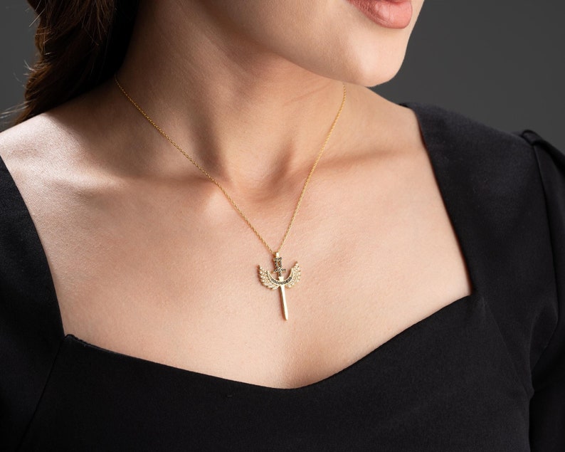 Puede incluir: Un collar de oro con un colgante que representa una espada con alas. El colgante est&aacute; decorado con peque&ntilde;as piedras.