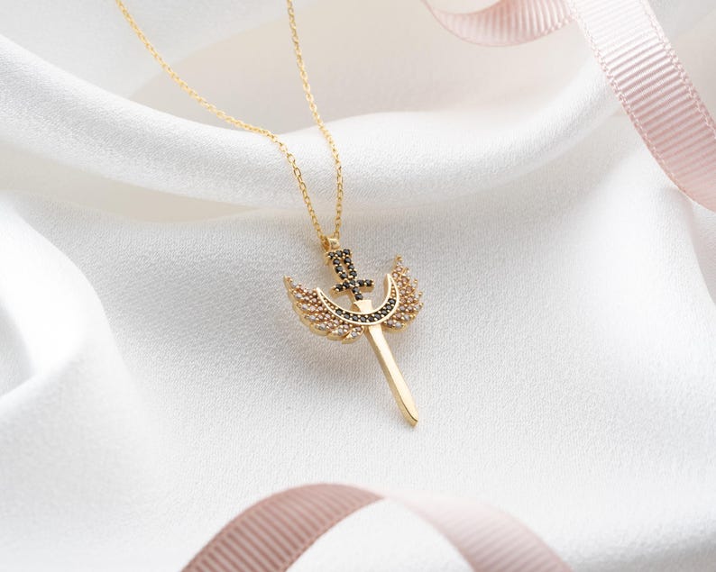 Puede incluir: Un collar de oro con un colgante que representa una espada con alas y una luna creciente. El colgante est&aacute; adornado con cristales negros y blancos.