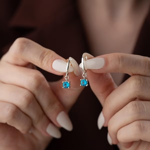Könnte beinhalten: Ein Paar silberne Creolen mit einem kleinen, quadratischen, blauen Edelstein, der an jeder Creole baumelt.