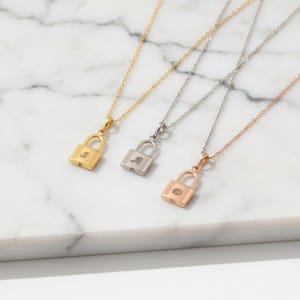 Puede incluir: Tres collares personalizados con forma de candado en oro, plata y oro rosa. Cada colgante tiene una inicial diferente: "S", "A" y "D". Los collares se exhiben sobre una superficie de mármol blanco, mostrando las delicadas cadenas y colgantes.