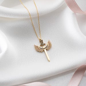 Puede incluir: Un collar de oro con un colgante que representa una espada con alas. La espada y las alas est&aacute;n adornadas con piedras brillantes negras y transparentes.