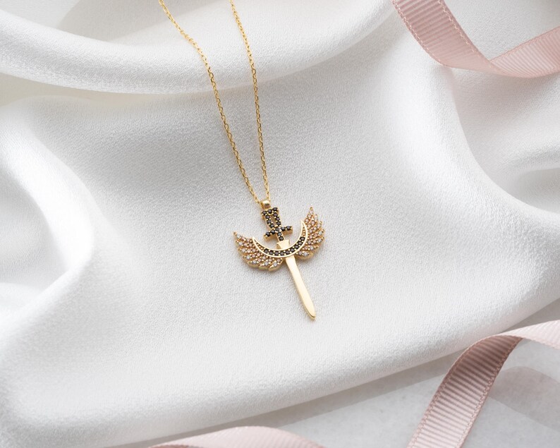 Puede incluir: Un collar de oro con un colgante que representa una espada con alas. La espada y las alas est&aacute;n adornadas con piedras brillantes negras y transparentes.