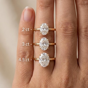 May include: Three gold rings with oval-cut clear stones in varying sizes. The rings are labeled with text indicating weights of 2 ct, 3 ct, and 4.5 ct. Each ring features a thin, polished gold band and a prong setting holding the central stone.