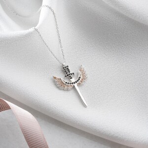 Puede incluir: Un collar de plata con un colgante que representa una espada con alas. Las alas est&aacute;n decoradas con cristales transparentes y negros. La espada tiene un cristal negro en la parte superior.