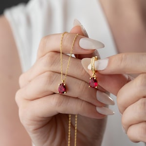 May include: A gold necklace and earrings set with a red gemstone pendant. The necklace has a delicate chain and the earrings are small and simple.