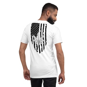 Op de afbeelding: Een wit T-shirt met een zwart-wit Amerikaans vlagontwerp met een marihuanablad. De vlag is verweerd en heeft een grunge-look.