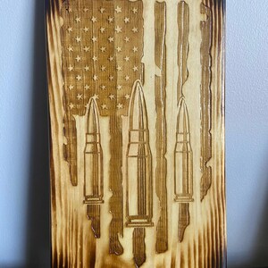 May include: Wooden plaque with a carved American flag design and three bullet casings. The wood has a natural grain with a dark, burnt border. The flag features stars and stripes, with the bullet casings positioned below.