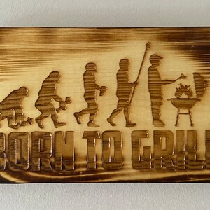 May include: A wooden sign with a burnt wood finish features a silhouette of the evolution of man, culminating in a person grilling. The text "BORN TO GRILL" is below the silhouettes.