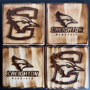 May include: Four wooden coasters with a burnt wood finish. Each coaster features a Creighton Bluejays logo with a bird mascot. Two coasters have the full logo with the text "CREIGHTON BLUEJAYS" and two coasters have just the bird mascot.