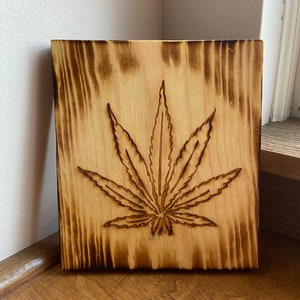 May include: A wood burned wall hanging with a cannabis leaf design. The wood is a light brown color with dark brown wood grain.
