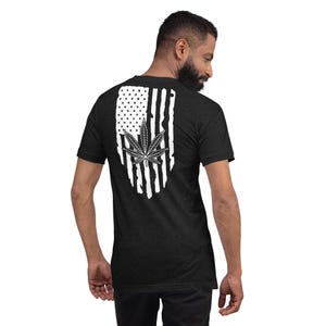 Op de afbeelding: Een zwart T-shirt met een wit, verweerd Amerikaans vlagontwerp met een marihuanablad in het midden.