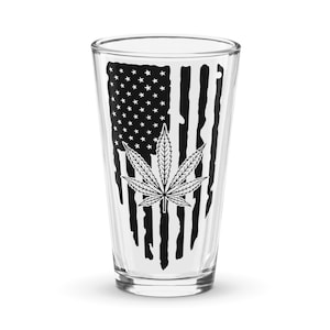 Op de afbeelding: Een helder glazen pintglas met een zwart-wit ontwerp van een marihuanablad en een Amerikaanse vlag. De vlag is verweerd en heeft een vervaagde uitstraling.