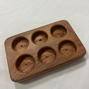 May include: A rectangular wooden shot glass holder with six circular indentations. The holder is made of a light brown wood and has a smooth finish. Each indentation has a small hole in the centre.