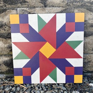 May include: A barn quilt with a geometric design featuring a red, yellow, green, and purple color scheme. The quilt is made of wood and has a white background.