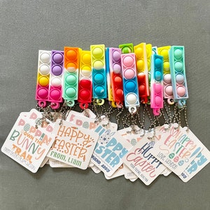 May include: A collection of colorful rectangular fidget toys with attached keychains. Each toy features multiple bubble-like circles in various colors. Attached are tags with Easter-themed text and graphics, including phrases like "Happy Easter" and "Poppin' Down the Bunny Trail."