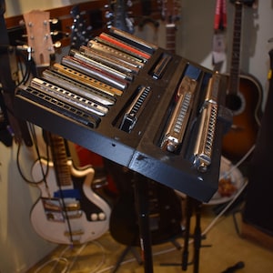 Puede incluir: Un soporte de exhibición negro que contiene una colección de armónicas. Las armónicas son plateadas y doradas, con varios diseños. Se ven guitarras en el fondo, lo que sugiere un tema musical.