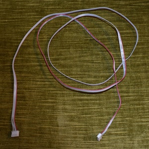 May include: A white cable with red wires running through it. The cable has two white connectors on the ends. The cable is coiled up on a green surface.