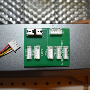 Könnte beinhalten: Eine grüne Leiterplatte mit weißen Steckverbindern, die mit "Axis", "Ortur/Neje/TwoTrees", "Atomstack", "XTool", "Laser", "XTool" und "Other" beschriftet sind. Ein weißer Stecker mit vier Drähten ist an die Platine angeschlossen.