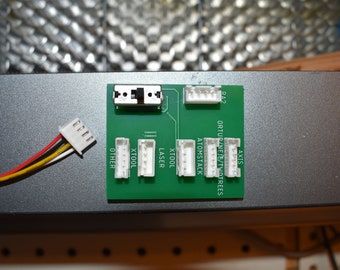 Pode incluir: Uma placa de circuito verde com conectores brancos etiquetados como "Axis", "Ortur/Neje/TwoTrees", "Atomstack", "XTool", "Laser", "XTool" e "Other". Um conector branco com quatro fios está conectado à placa.