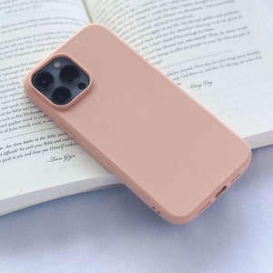 May include: A light pink phone case rests on an open book. The phone case has a smooth, matte finish and a camera module with multiple lenses. The book's pages are white with black text. The scene is lit with soft, natural light.