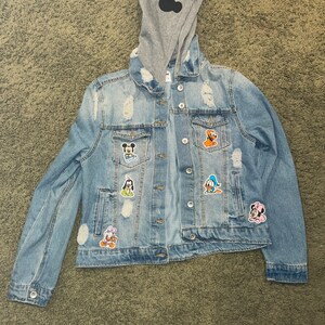 May include: A light blue denim jacket with a grey hooded sweatshirt underneath. The jacket has several patches of Disney characters, including Mickey Mouse, Donald Duck, Goofy, and Pluto.