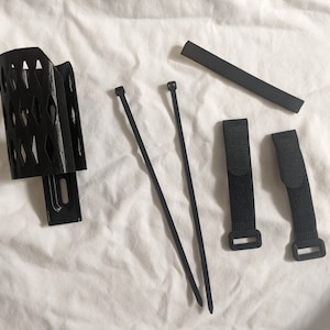 May include: A black plastic bike water bottle holder with a black plastic mounting bracket, two black cable ties, and two black hook and loop straps.