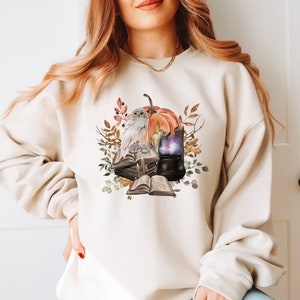 Könnte beinhalten: Beiges Sweatshirt mit einem Aquarelldesign einer weißen Eule, einem Kürbis, Büchern und einem Kessel. Das Design umfasst Herbstblätter und eine brennende Kerze, die ein Herbst- oder Halloween-Thema erzeugt. Das Sweatshirt hat eine neutrale Farbe.