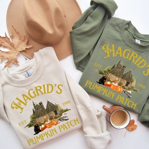 Könnte beinhalten: Zwei cremefarbene und grüne Sweatshirts mit einem grafischen Design eines Hauses mit einem Kürbisfeld und dem Text "Hagrid's Pumpkin Patch Est. 1980".