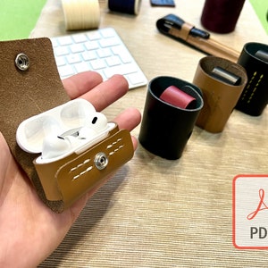 以下が含まれることがあります： スナップ留め具付きの茶色の革ケースで、ワイヤレスイヤホンを収納するように設計されています。ケースは開いており、イヤホンがケースの中にあります。ケースは、他の革ケースと一緒にテーブルの上に置かれています。