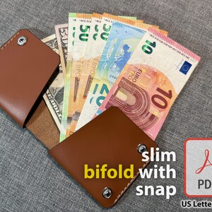 May include: A brown leather slim bifold wallet with snap closure, open to display various currencies, including US dollars and Euro banknotes. The wallet is shown on a gray surface. Text on the image reads "slim bifold with snap" and "PDF".