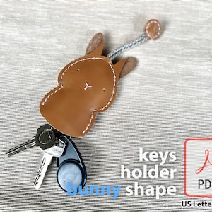 May include: Brown leather key holder shaped like a bunny with a white stitched outline. The key holder has a loop for attaching keys and a small brown leather circle at the end of a gray cord. The text "keys holder bunny shape" is written below the key holder. The text "PDF" is written in a red box with a white Adobe Acrobat logo. The text "US Letter & A4" is written below the box.