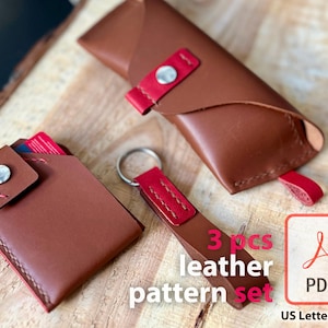 Puede incluir: Un conjunto de tres accesorios de cuero marrón: un tarjetero, un estuche para gafas y un llavero. Cada artículo presenta detalles rojos y un cierre de botón a presión plateado. Los artículos se muestran sobre una superficie de madera.